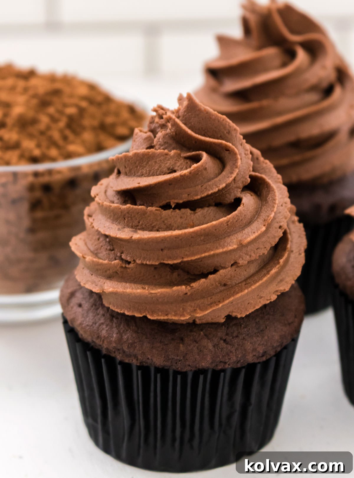 A beautifully frosted chocolate cupcake, crowned with a perfect swirl of the Best Chocolate Mint Buttercream Frosting, positioned invitingly next to a glass bowl filled with rich cocoa powder, emphasizing the delectable chocolate component.