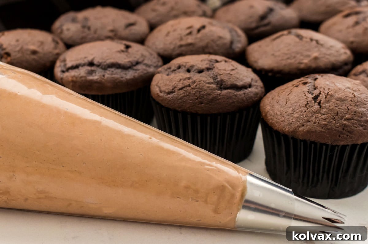 Closeup on a decorating bag filled with Mocha Frosting sitting in front of a batch of unfrosted chocolate cupcakes.