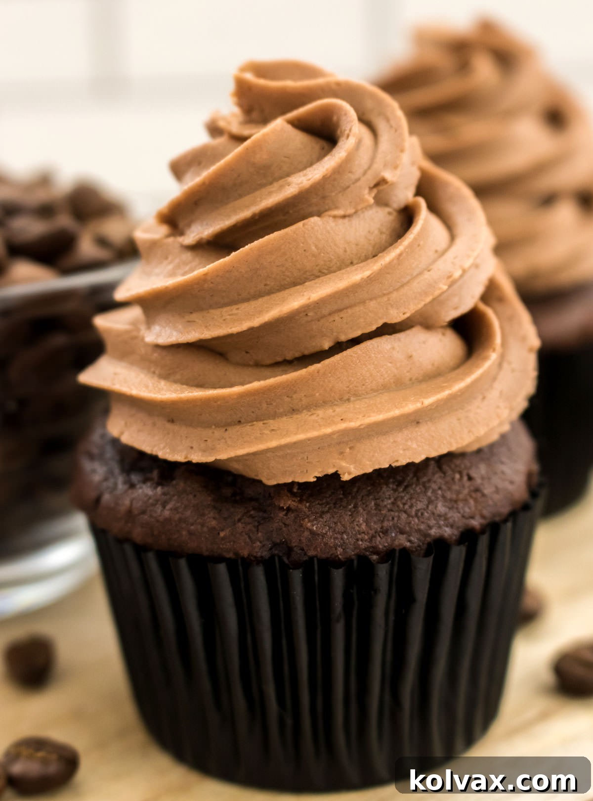 Closeup on a chocolate cupcake topped with The Best Mocha Buttercream Frosting sitting on a cutting board next to a glass bowl filled with coffee beans.