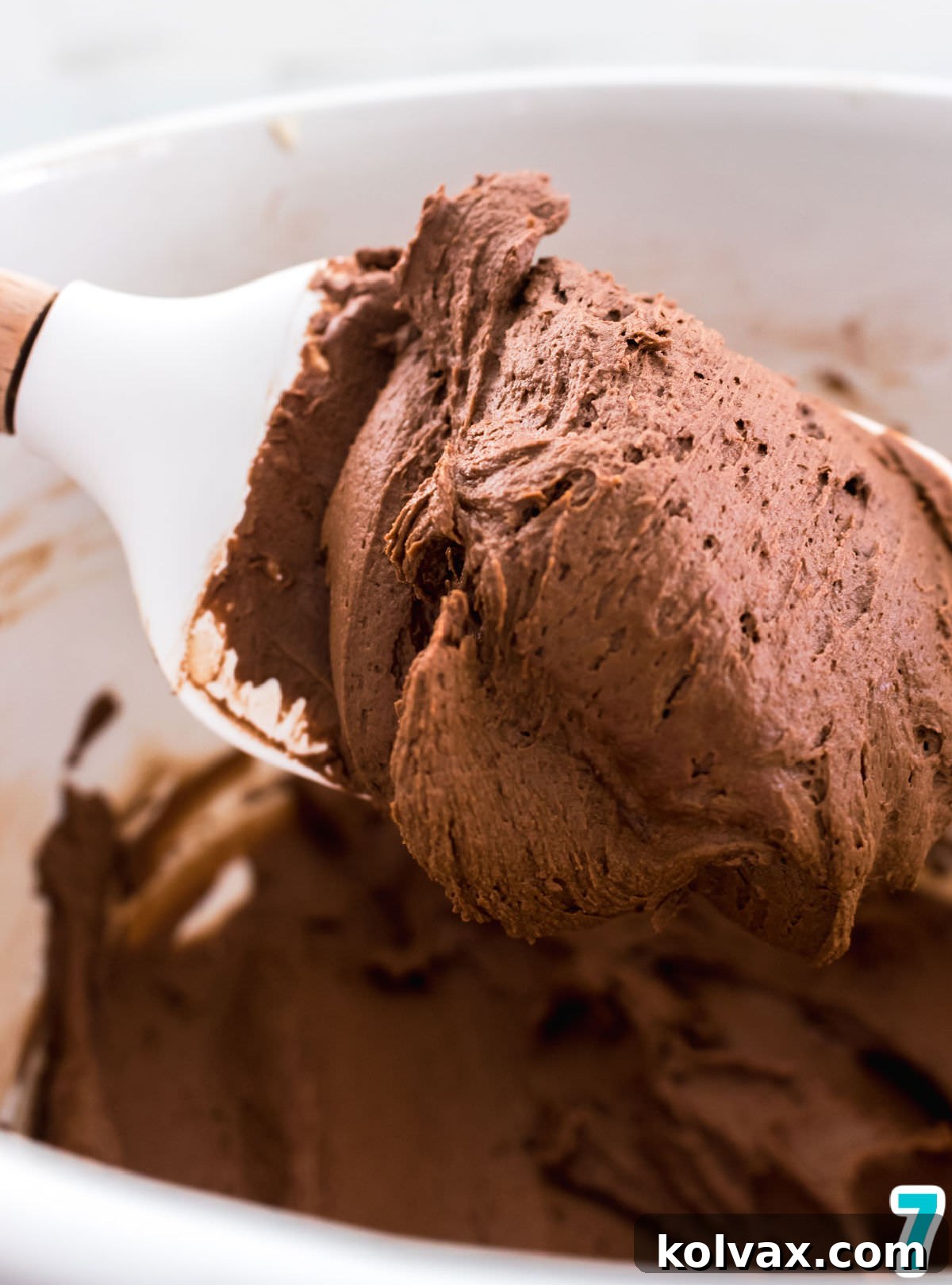 Closeup on a white mixing bowl filled with chocolate frosting and a white spatula.