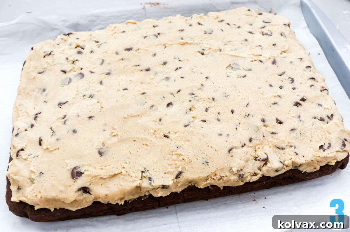 A baked chocolate cookie layer on parchment paper, gently topped with a generous layer of edible cookie dough frosting, ready for the final chocolate coating.