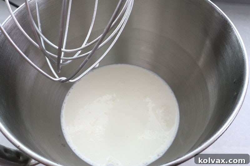 Pouring heavy whipping cream into a well-chilled stainless steel mixing bowl, a crucial step for achieving the perfect consistency for whipped cream frosting.