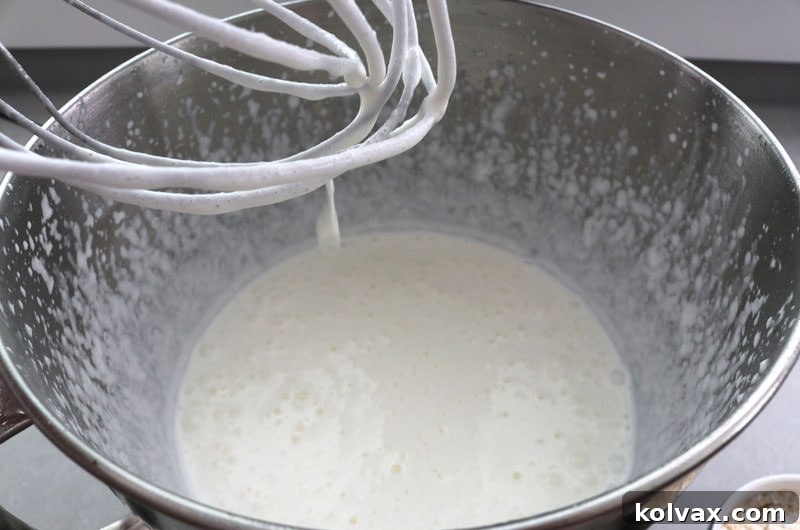 Electric mixer whipping heavy cream in a bowl, forming soft peaks. This stage is important before adding other ingredients to create a light and airy frosting base.