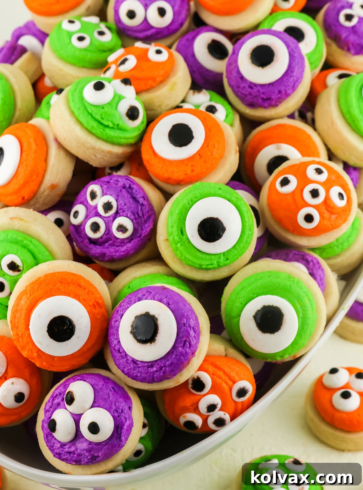 A vibrant collection of Mini Monster Sugar Cookies, each uniquely decorated with colorful frosting and playful candy eyes, artfully arranged in a white server bowl.