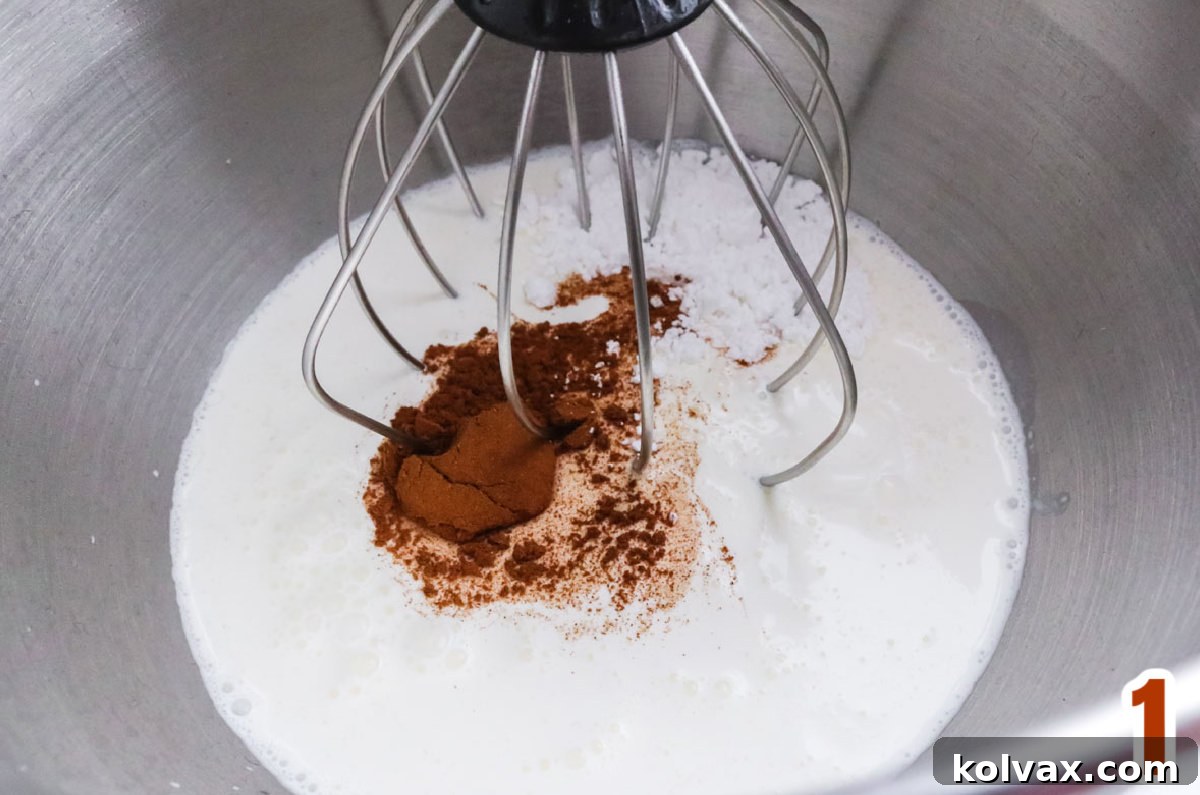 A vibrant close-up shot of a metal mixing bowl filled with the key ingredients for Homemade Cinnamon Whipped Cream: rich Heavy Whipping Cream, fine Powdered Sugar, and aromatic Ground Cinnamon.