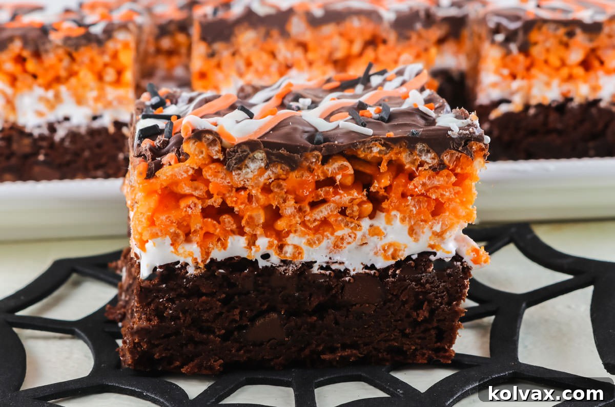 Close-up of a perfectly cut Halloween Brownie Rice Krispie Treat, featuring visible layers of brownie, marshmallow, orange Rice Krispie, and chocolate, resting on a decorative spider web. A platter of more treats is blurred in the background.