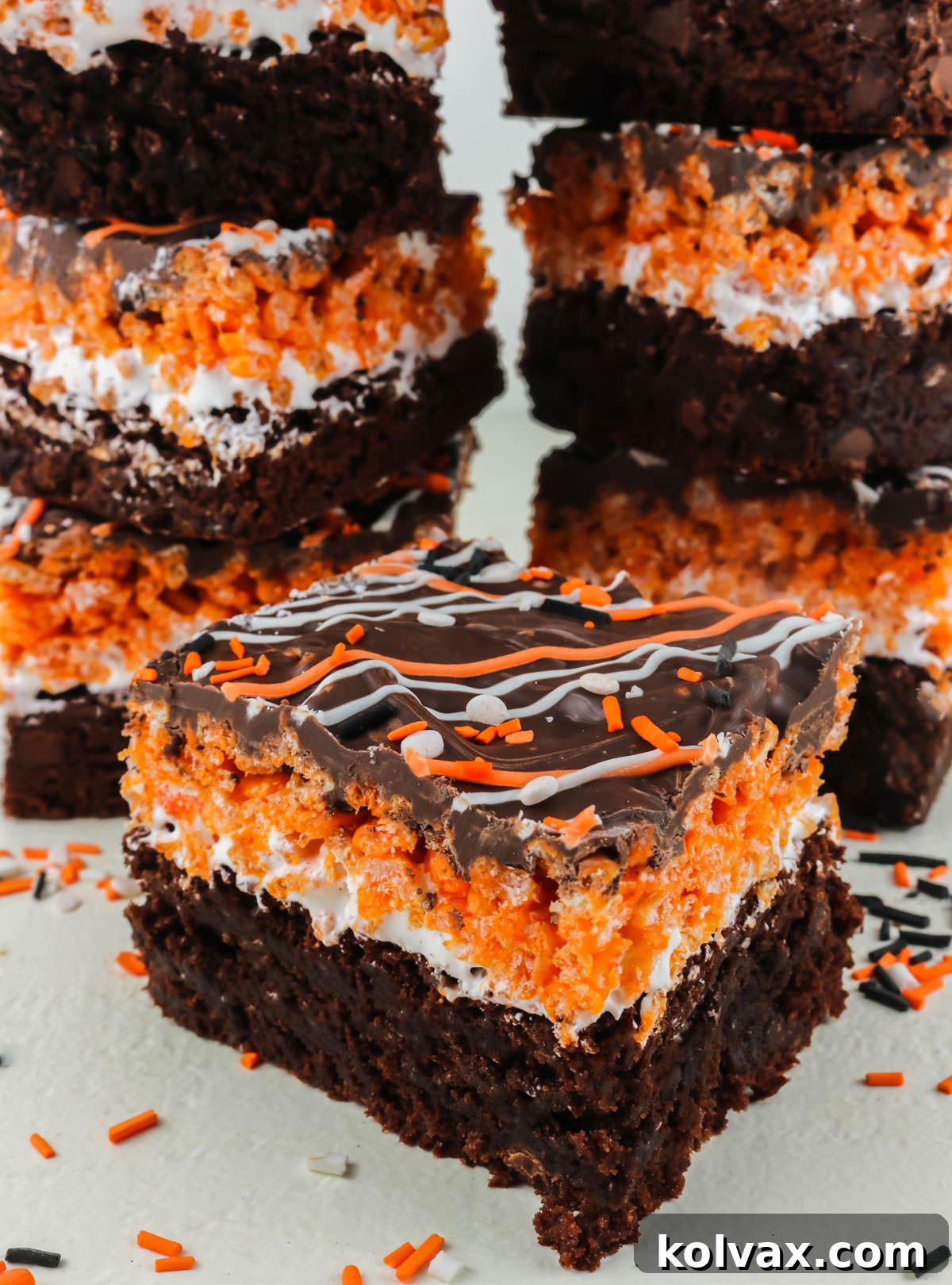 A close-up shot of a single Halloween Brownie Rice Krispie Treat, showcasing its distinct layers, with several other neatly stacked bars blurred in the background.