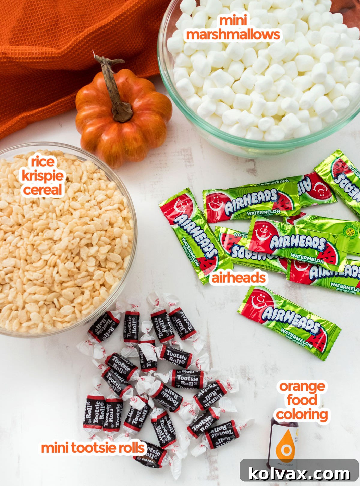 A vibrant flat lay of all the essential ingredients for making Pumpkin Rice Krispie Treats: a bowl of fluffy mini marshmallows, a box of crispy Rice Krispie cereal, a stick of butter, a bottle of concentrated orange food coloring, a pile of brown Mini Tootsie Rolls, and a selection of green Airhead candies, all artfully arranged on a clean white surface.