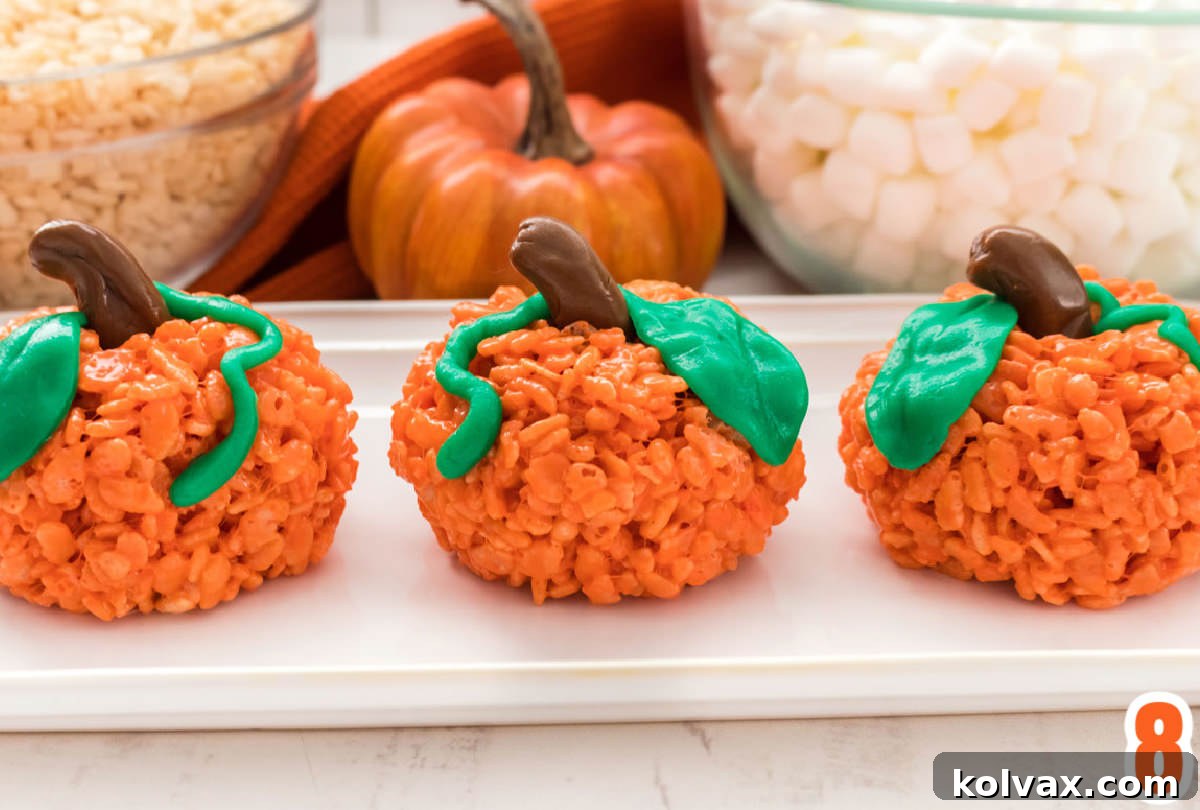 Three perfectly crafted Pumpkin Rice Krispie Treats nestled on a pristine white serving platter, positioned on a white table. A decorative pumpkin and the recipe ingredients, now in the background, complete the festive fall tableau, ready for display or enjoyment.