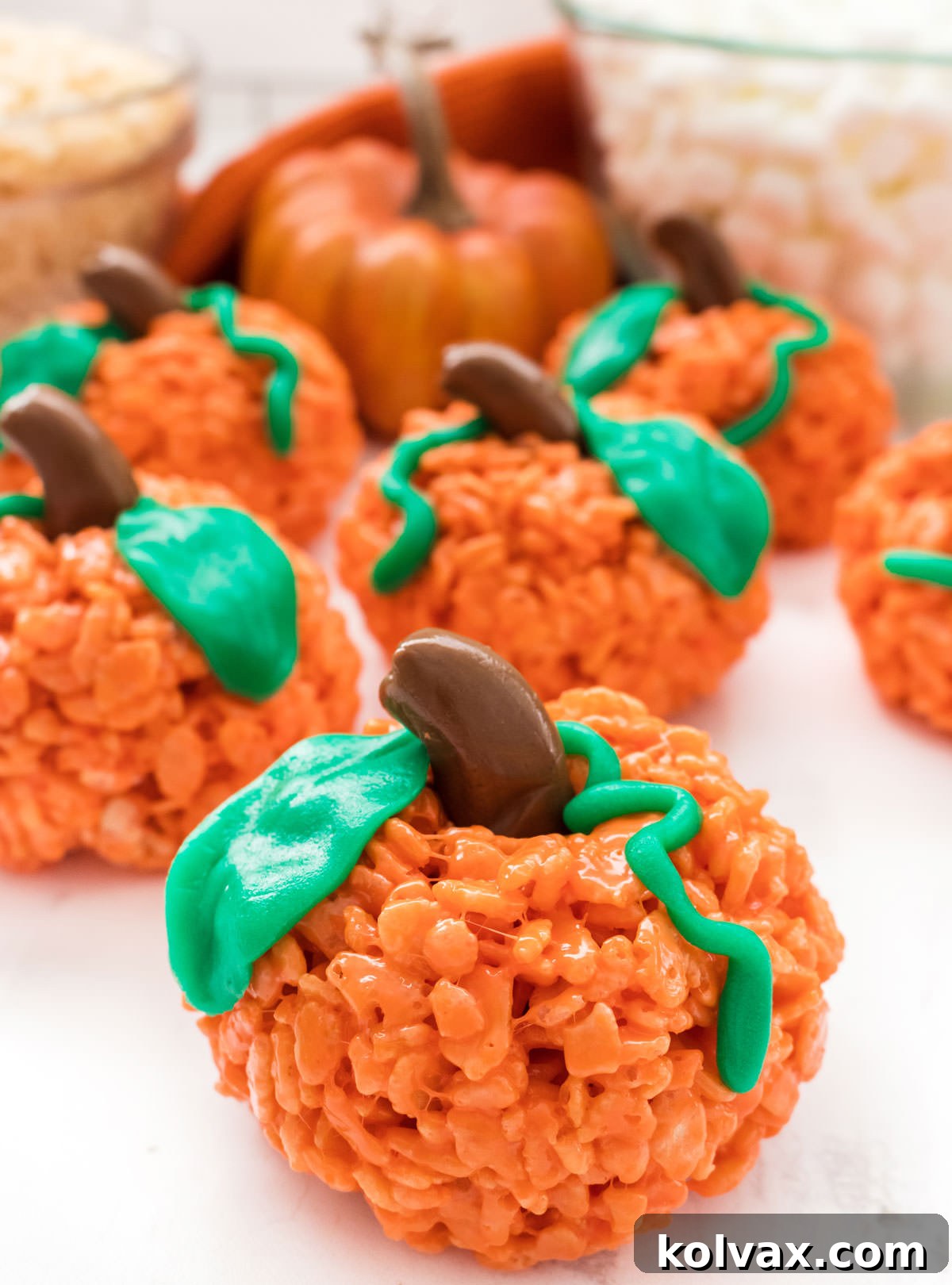 A beautiful close-up shot of six completed Pumpkin Rice Krispie Treats, artfully arranged on a white surface. A decorative pumpkin, a bowl of mini marshmallows, and a bowl of Rice Krispie cereal are softly blurred in the background, creating a warm and inviting fall-themed setting.