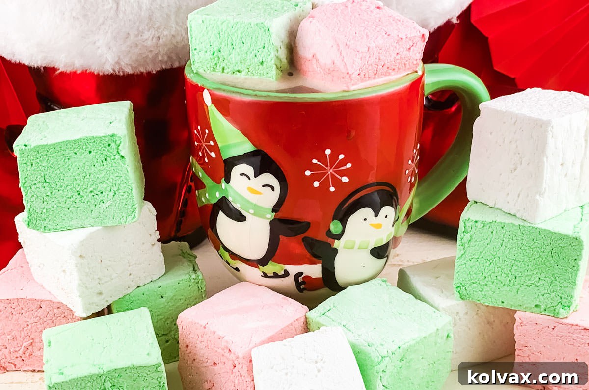 Red, Green and White Christmas Homemade Marshmallows sitting on a white surface in front of a Christmas Coffee Mug filled with hot chocolate and two homemade marshmallows.