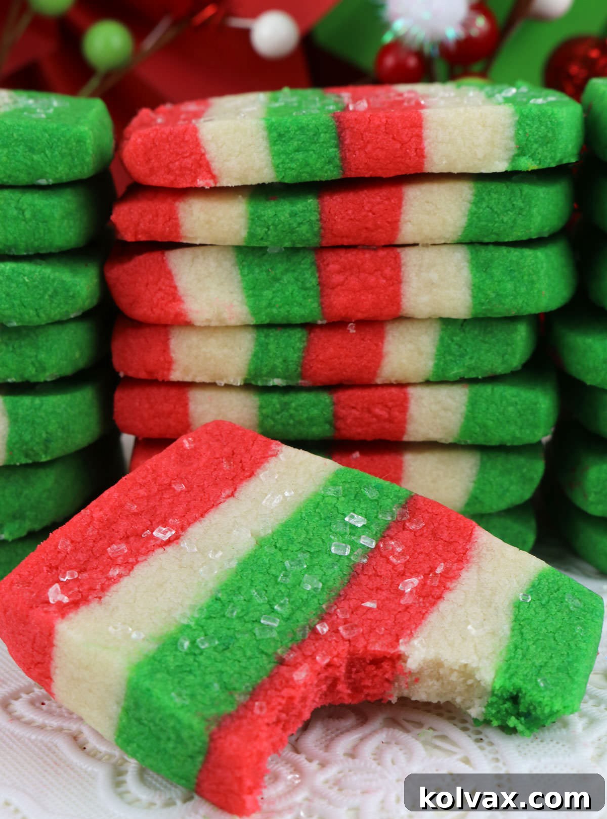A close-up of a delightful Christmas Butter Cookie with a bite taken out, resting in front of a tall, festive stack of red, white, and green cookies, all adorned with sparkling sugar.