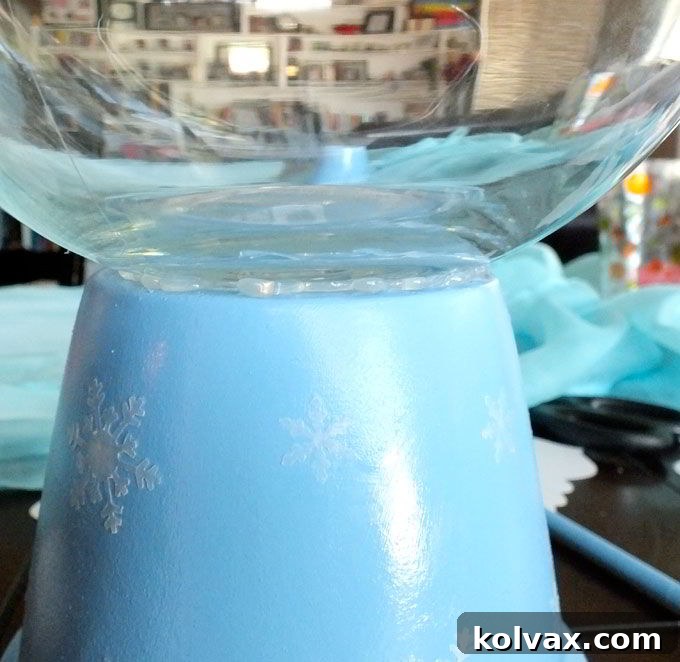 Using hot glue to securely attach the glass fishbowl vase to the base of the painted terra cotta pot, forming the core of your Disney Frozen Centerpiece.