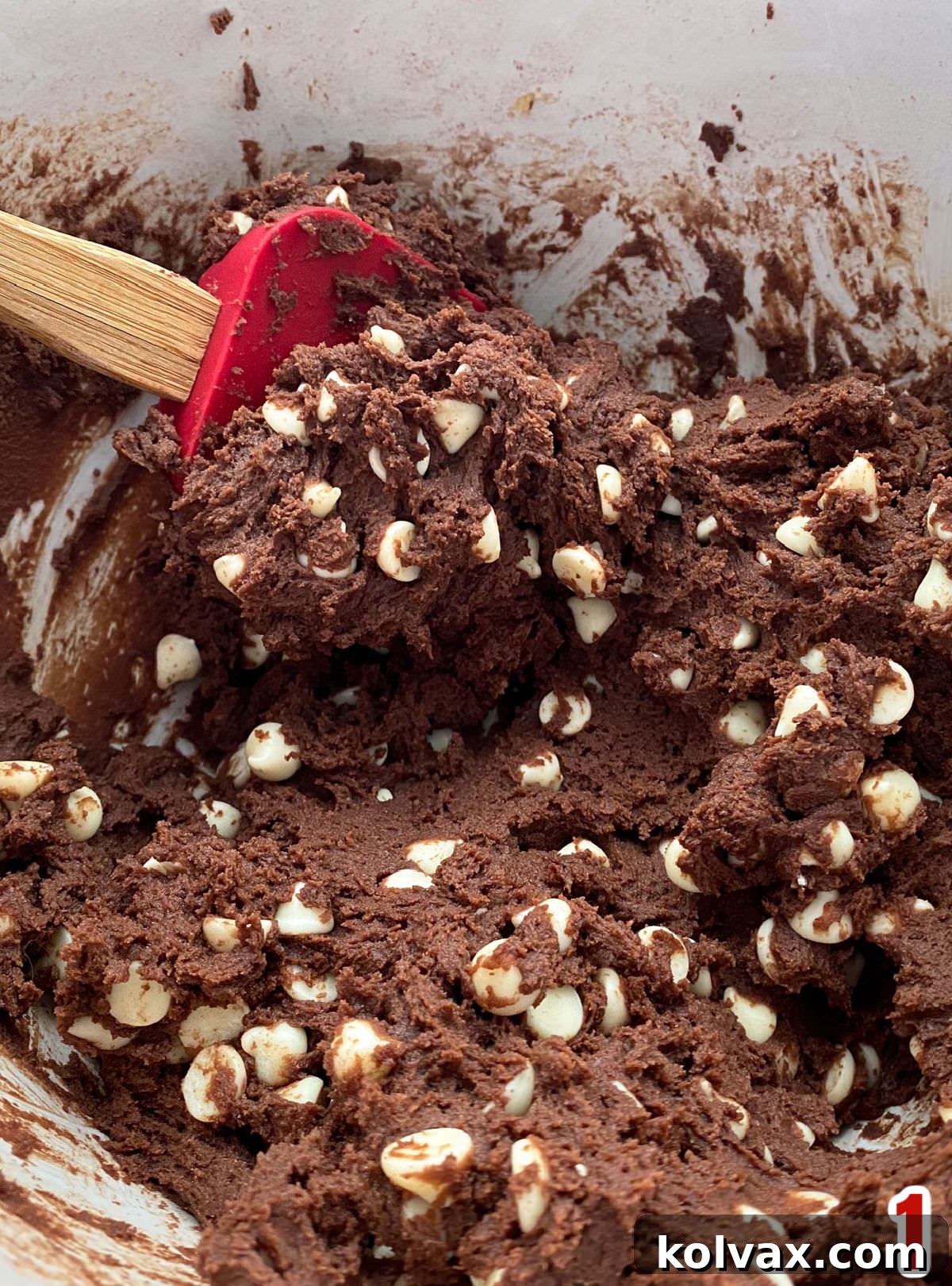 Closeup on a white mixing bowl filled with rich, dark Chocolate White Chocolate Chip Cookie Dough, ready for baking, with a wooden spatula resting nearby.