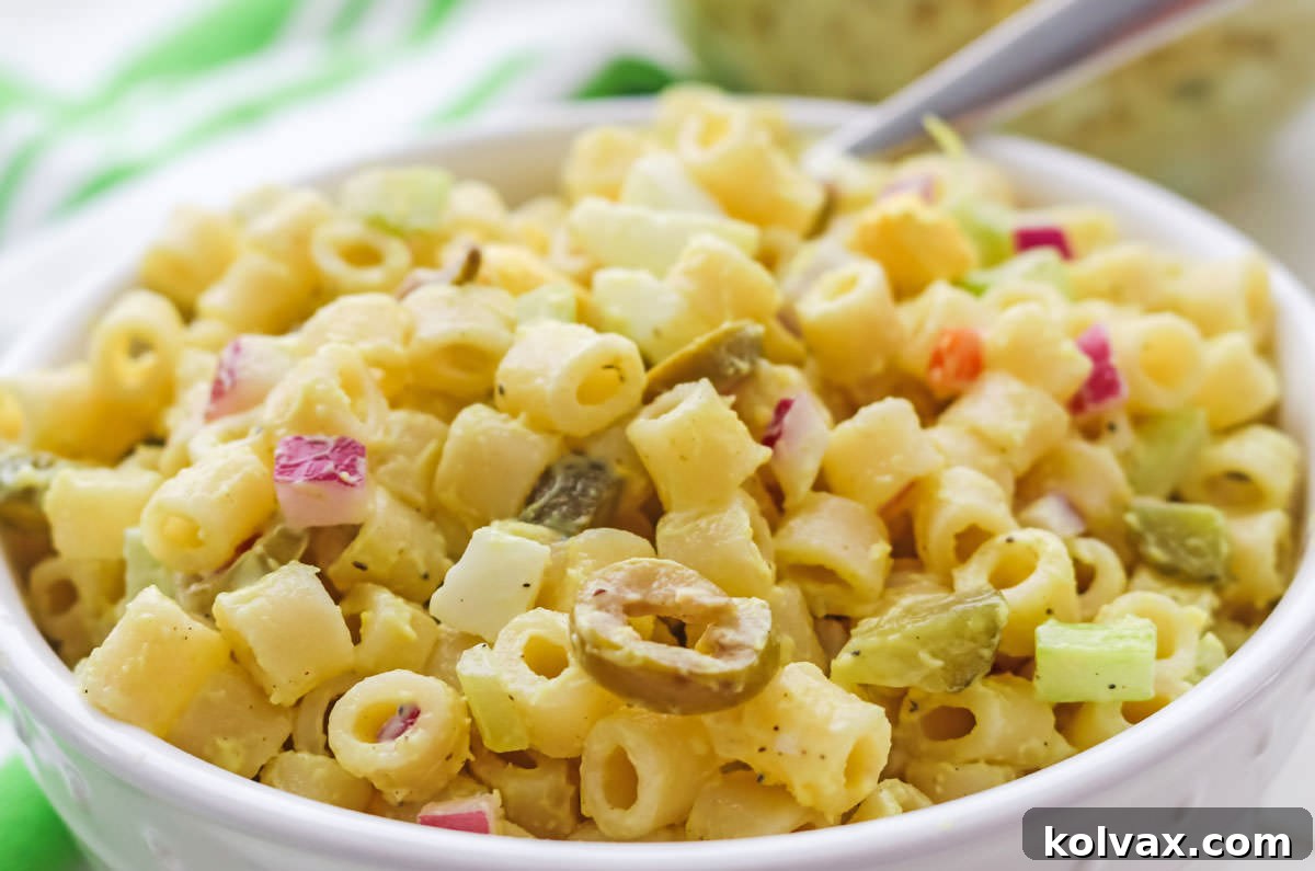 Closeup of a pristine white bowl generously filled with creamy Classic Macaroni Salad, featuring tender macaroni pasta, crisp vegetables, and a luscious dressing. A silver spoon is positioned in front of a soft green and white checkered towel, inviting a taste.