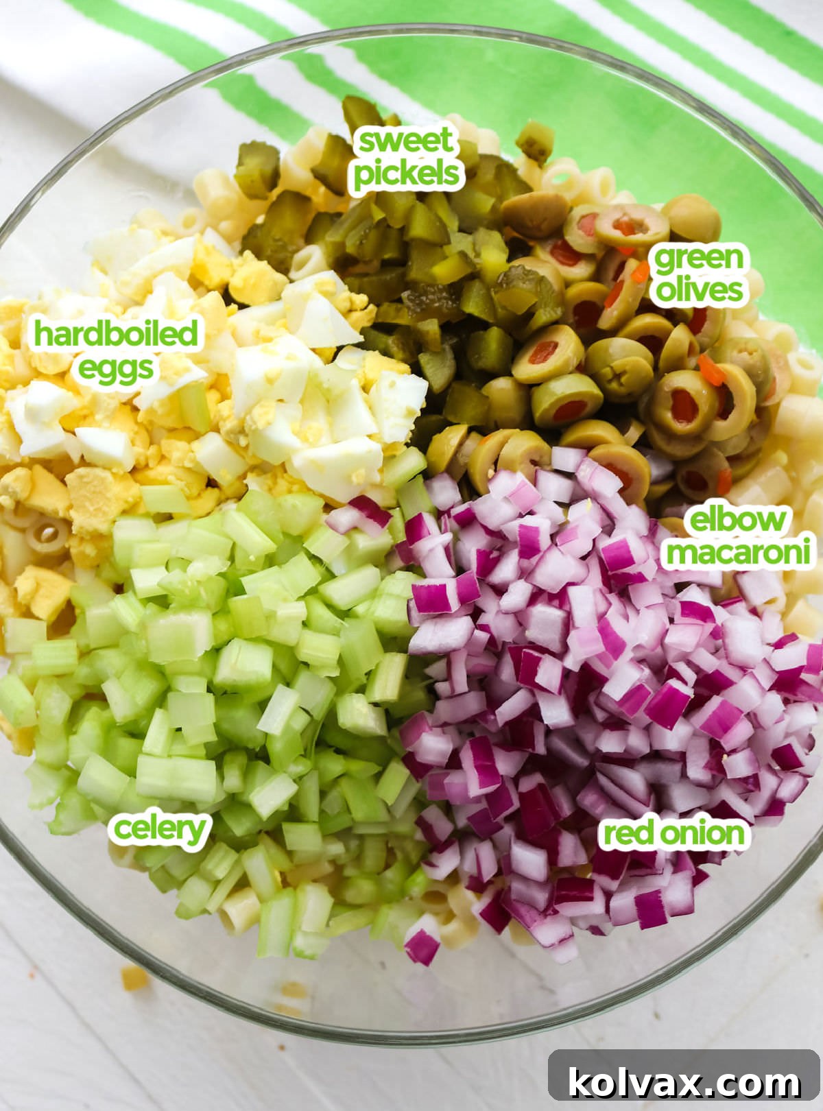 All the essential ingredients neatly arranged for making Classic Macaroni Salad. Displayed are short macaroni pasta, a jar of sweet pickles, vibrant green olives, a fresh red onion, crisp celery stalks, and perfectly hard-boiled eggs, ready for preparation.