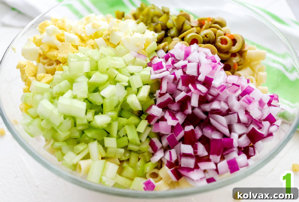 A clear glass mixing bowl holds the foundational ingredients for Classic Macaroni Salad: perfectly cooked short macaroni pasta, alongside freshly chopped green olives, finely diced red onion, crisp celery, and creamy hard-boiled eggs, all ready to be combined.