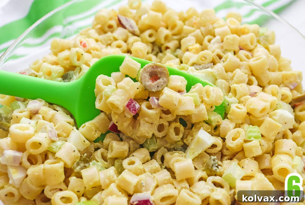 A close-up shot of a clear glass bowl generously filled with freshly made Classic Macaroni Salad. A green serving spoon is poised, scooping out a portion of the creamy, vegetable-packed pasta salad, highlighting its inviting texture.