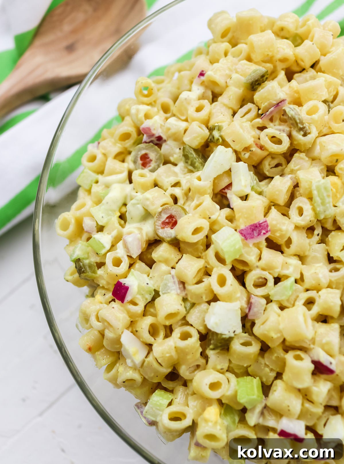 A beautifully presented clear glass serving bowl brimming with fresh Classic Macaroni Salad rests on a clean white table. In the background, a soft towel and a rustic wooden spoon add a touch of homey charm, inviting guests to serve themselves.