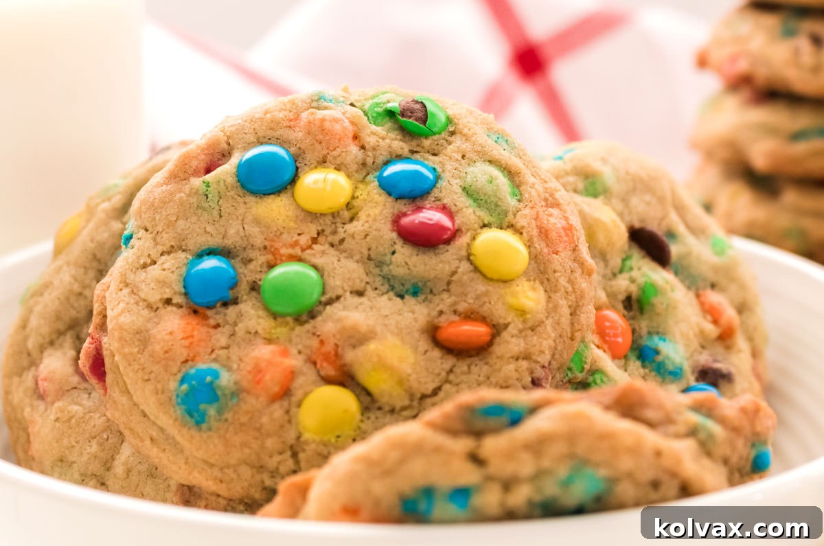 A close-up of a white bowl filled with five freshly baked Mini M&M Cookies, alongside a refreshing glass of milk and a stack of colorful cookies. The scene evokes comfort and deliciousness, highlighting the inviting texture and vibrant M&M candies.