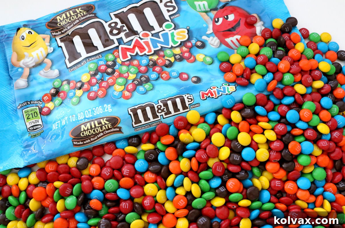 A close-up shot of a bag of M&M Minis spilling out onto a clean white table, showcasing their small size and vibrant colors. The image emphasizes the key ingredient for these delightful cookies.