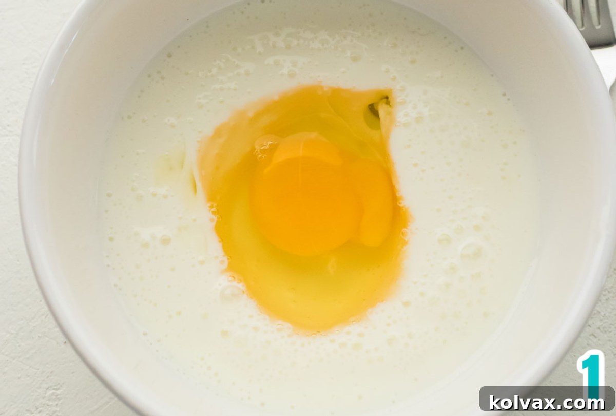 Closeup on a small white bowl filled with creamy buttermilk and a single cracked egg, ready to be combined.