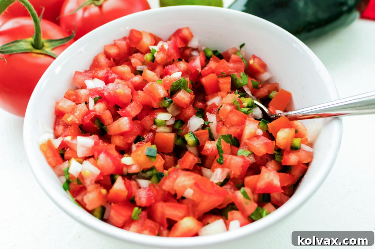 Closeup on a white bowl filled with Pico de Gallo sitting on a white table in front of tomatoes on the vine and a pablono pepper.