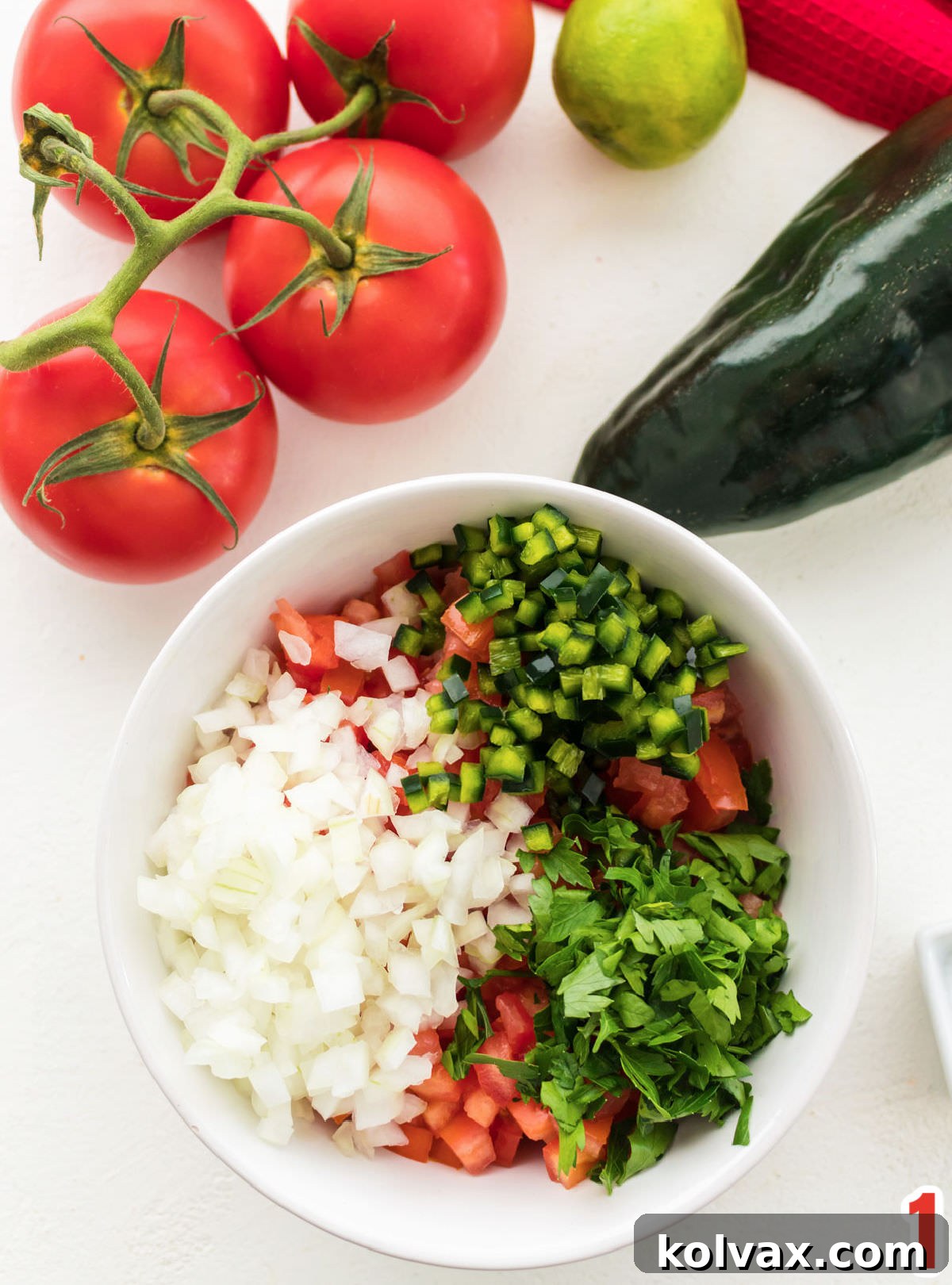 White bowl filled with diced tomatoes, onions, pablono peppers and cilantro sitting in front of tomatoes on the vine, lime and a pepper.