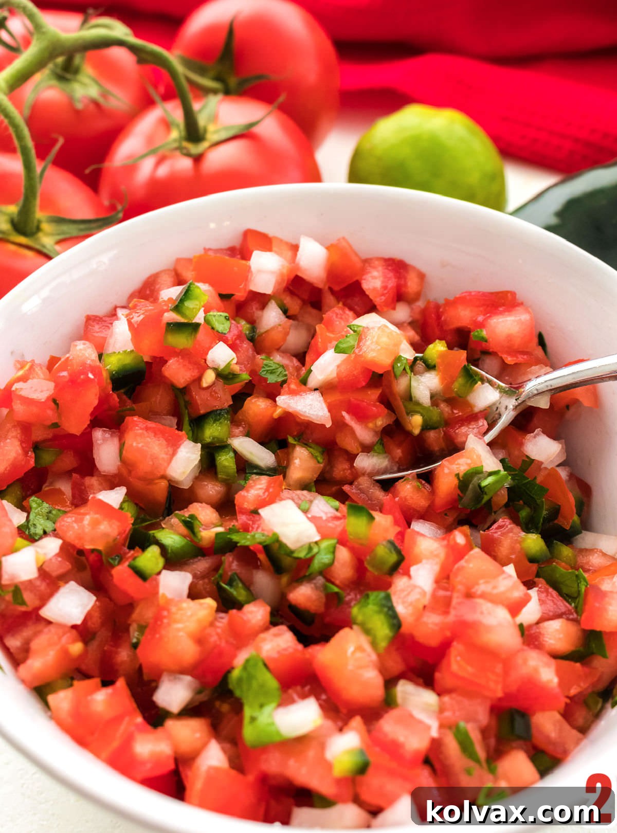 Closeup on a white serving bowl filled with Pico de Gallo with tomatoes, a lime and a pepper in the background.