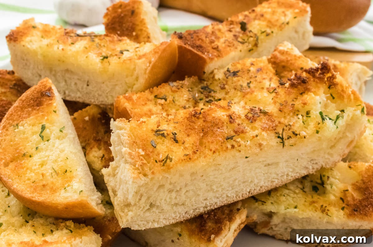 Closeup of golden brown homemade garlic bread slices stacked on a white plate, with a loaf of French bread and fresh garlic in the background.