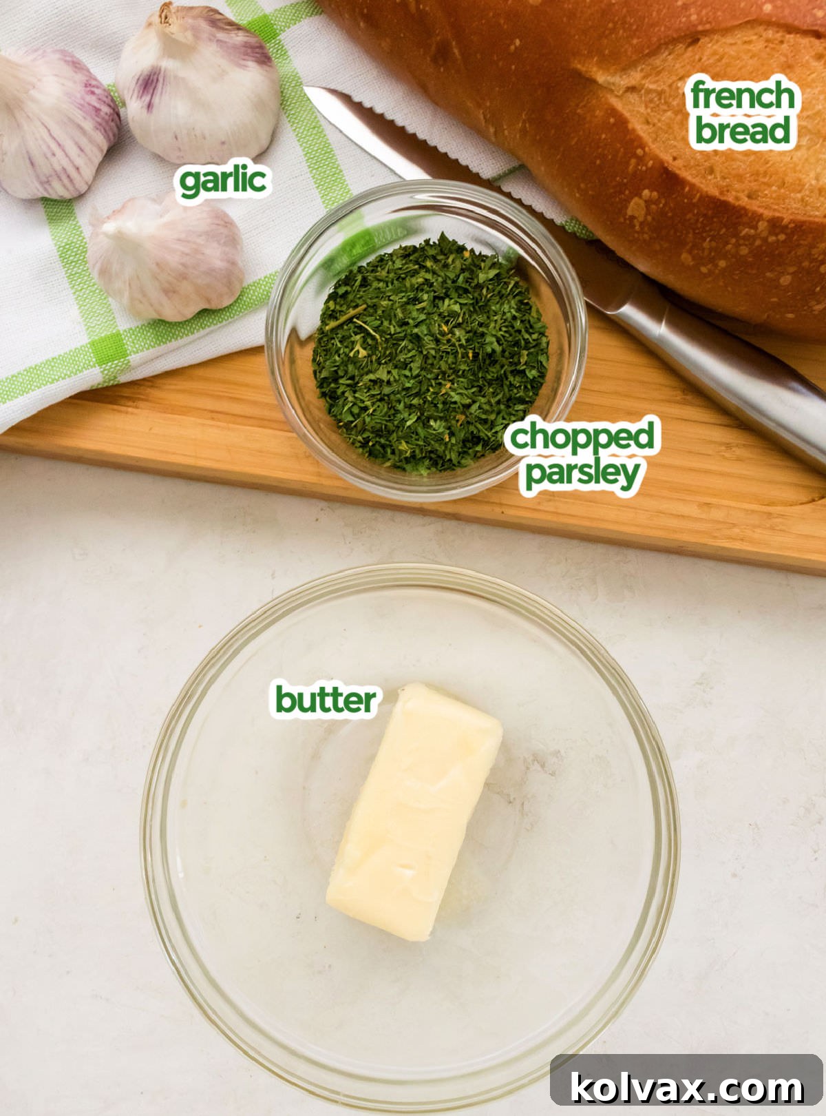 All the necessary ingredients laid out for making garlic bread: a loaf of French bread, fresh garlic cloves, a stick of butter, and a small bowl of chopped parsley.