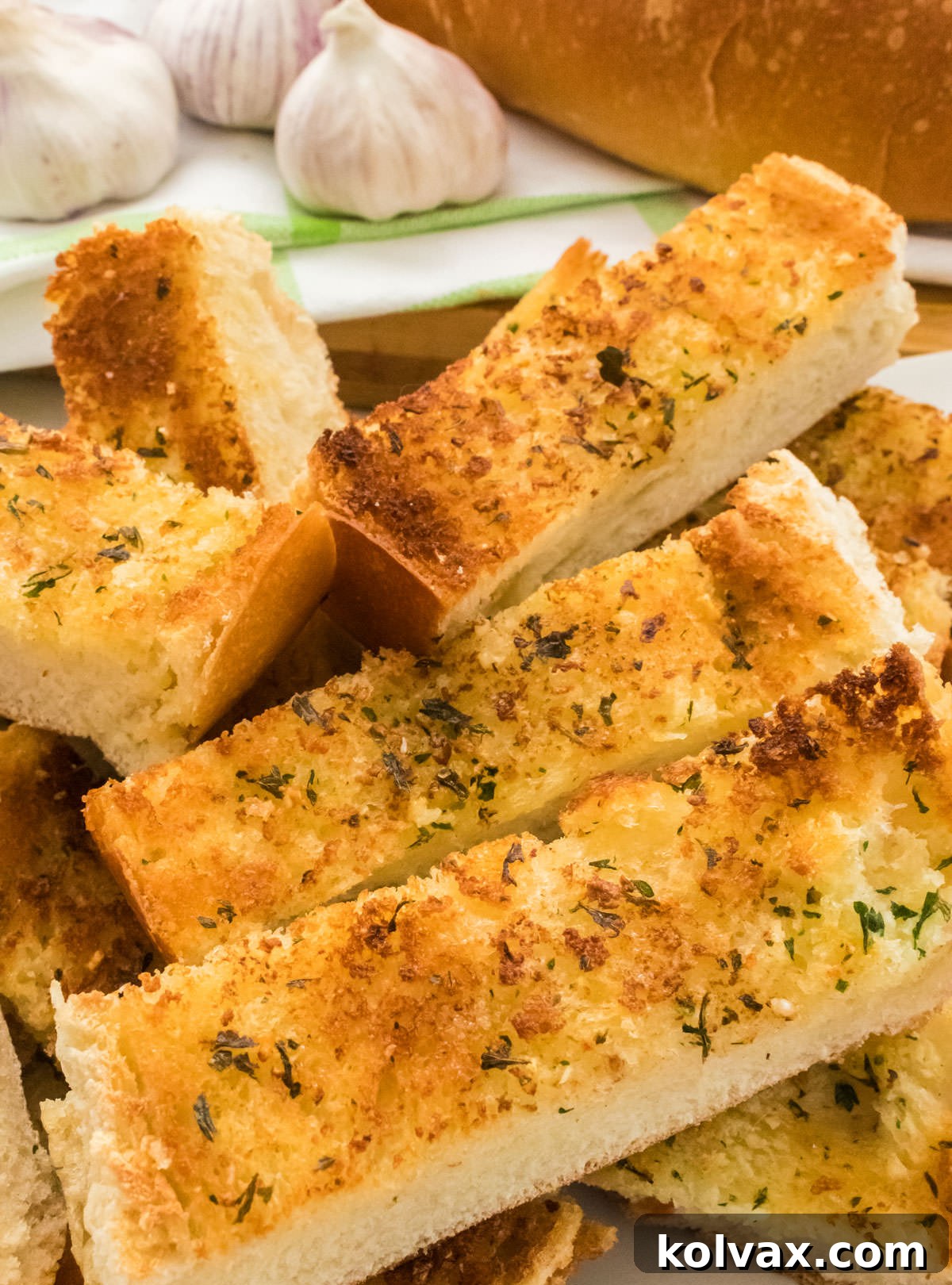 A beautiful stack of homemade garlic bread slices arranged on a white serving dish, with garlic bulbs and a French bread loaf in the background.