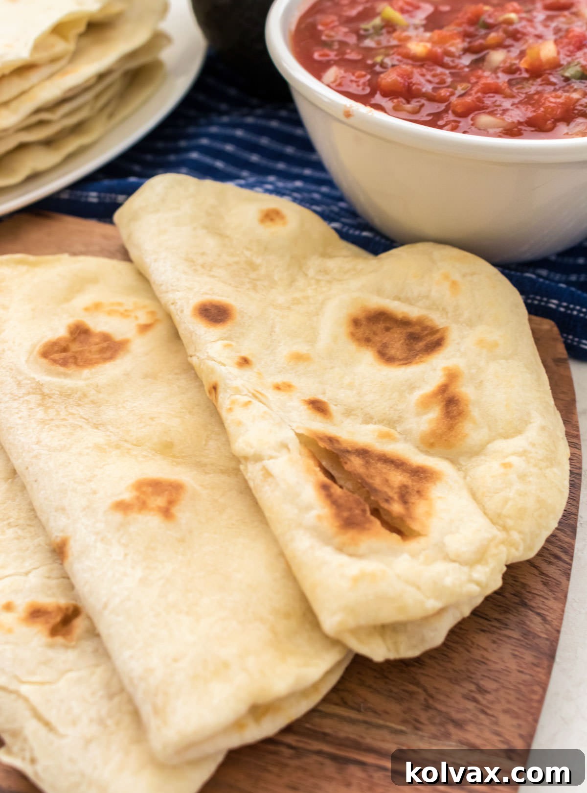 A close-up view of three soft homemade flour tortillas, folded in half, arranged on a cutting board, with a large stack of freshly cooked tortillas and a bowl of vibrant Pico de Gallo in the background.