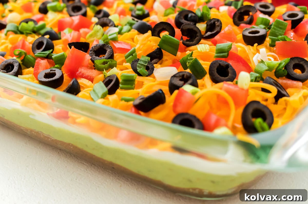 A vibrant, close-up shot of a classic 7 Layer Dip in a clear glass baking dish, showcasing distinct layers of refried beans, guacamole, sour cream, cheese, tomatoes, olives, and green onions. Perfect for parties and gatherings.