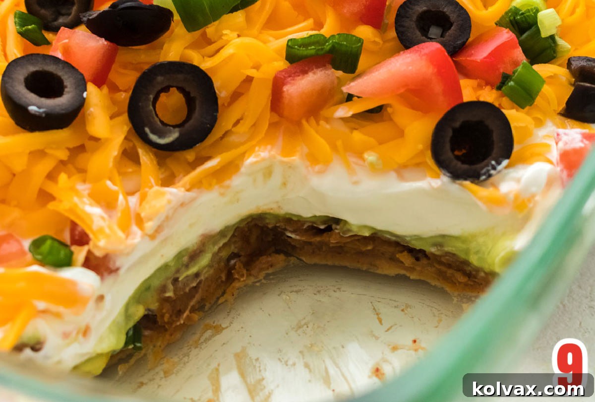 A close-up shot of a clear glass dish of 7 Layer Dip with a section scooped out, clearly revealing all seven distinct layers: refried beans, guacamole, sour cream, cheese, tomatoes, olives, and green onions. It highlights the perfect layering and inviting texture.