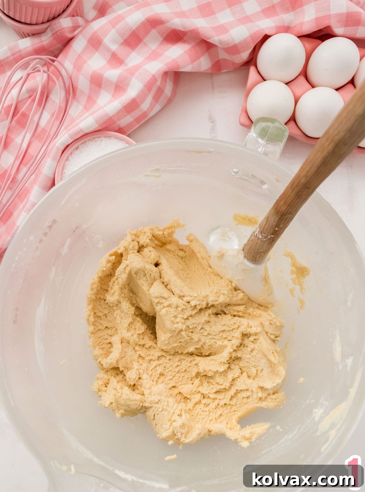 Closeup on a glass mixing bowl filled with Sugar Cookie dough and a white wood spatula on a white table surrounded by eggs, sugar and a pink table linen.