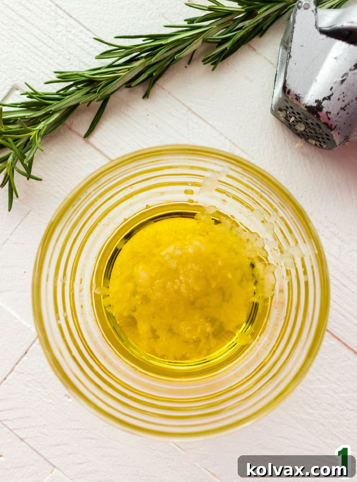 Closeup on a glass ramekin filled with olive oil and crushed garlic sitting on a white table next to a garlic press and a sprig of fresh rosemary.