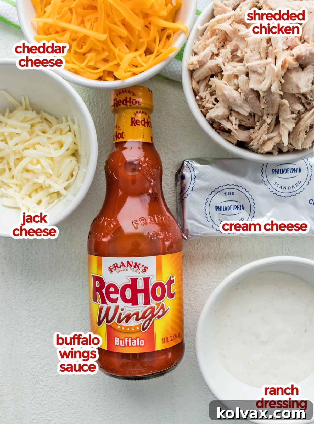 A top-down view of all the essential ingredients laid out for making Buffalo Chicken Dip, including a bowl of shredded chicken, blocks of cream cheese, a bottle of buffalo wings sauce, a bowl of ranch dressing, and piles of shredded cheddar and Monterey Jack cheeses.