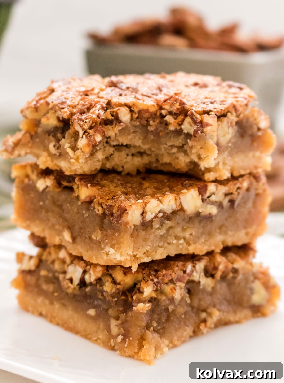 Closeup on a stack of three Pecan Pie Bars sitting on a white plate, with the top bar showing a delicious bite taken out, revealing the gooey filling and crunchy pecans within a tender crust.