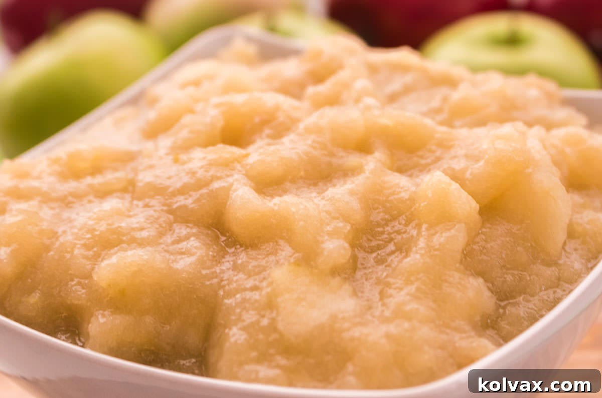 Closeup on a white bowl filled with The Best Homemade Applesauce sitting on a cutting board, garnished with a cinnamon stick and fresh apple slices.