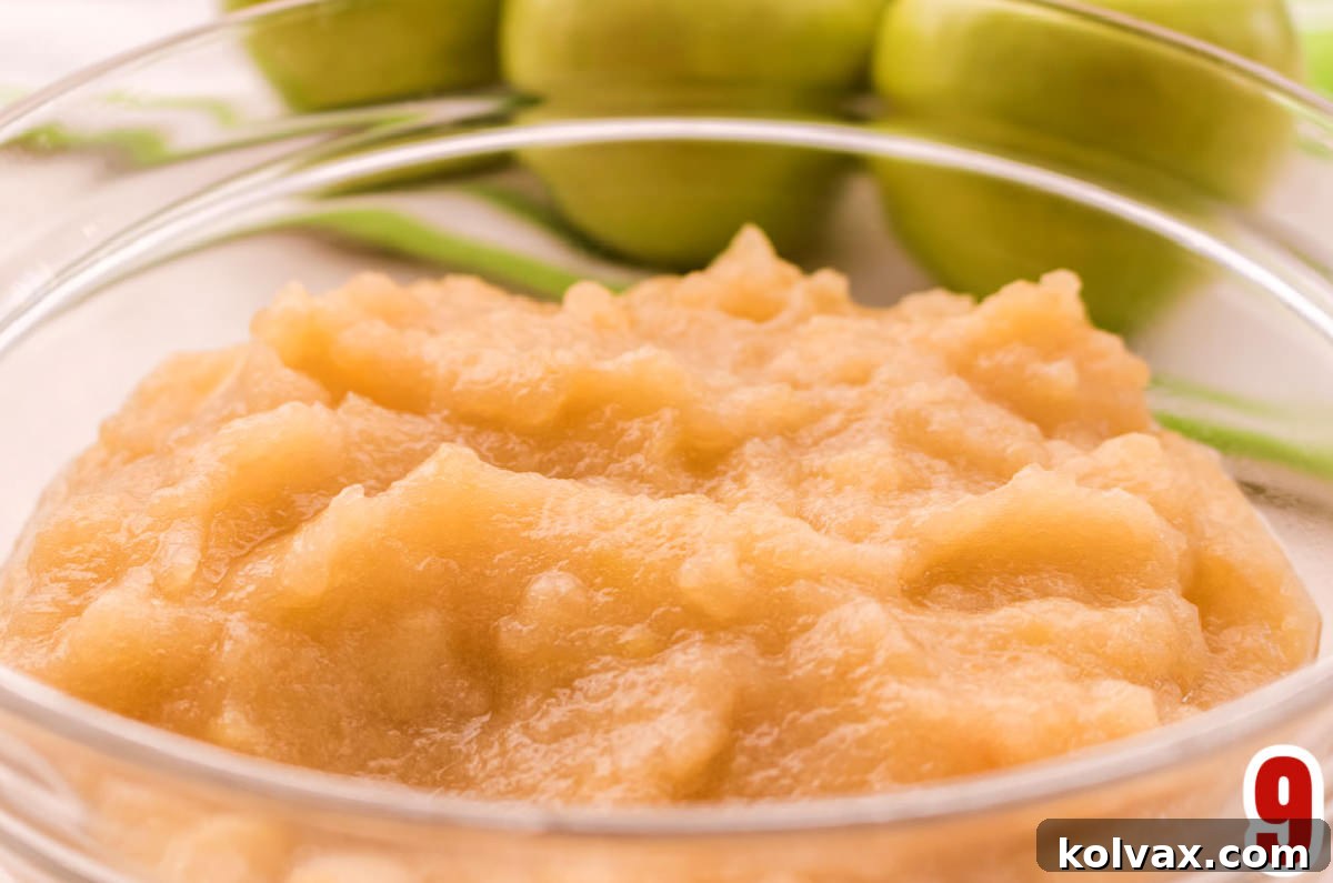 Closeup on a glass bowl filled with The Best Homemade Applesauce sitting in front of three green apples, garnished with a mint leaf.