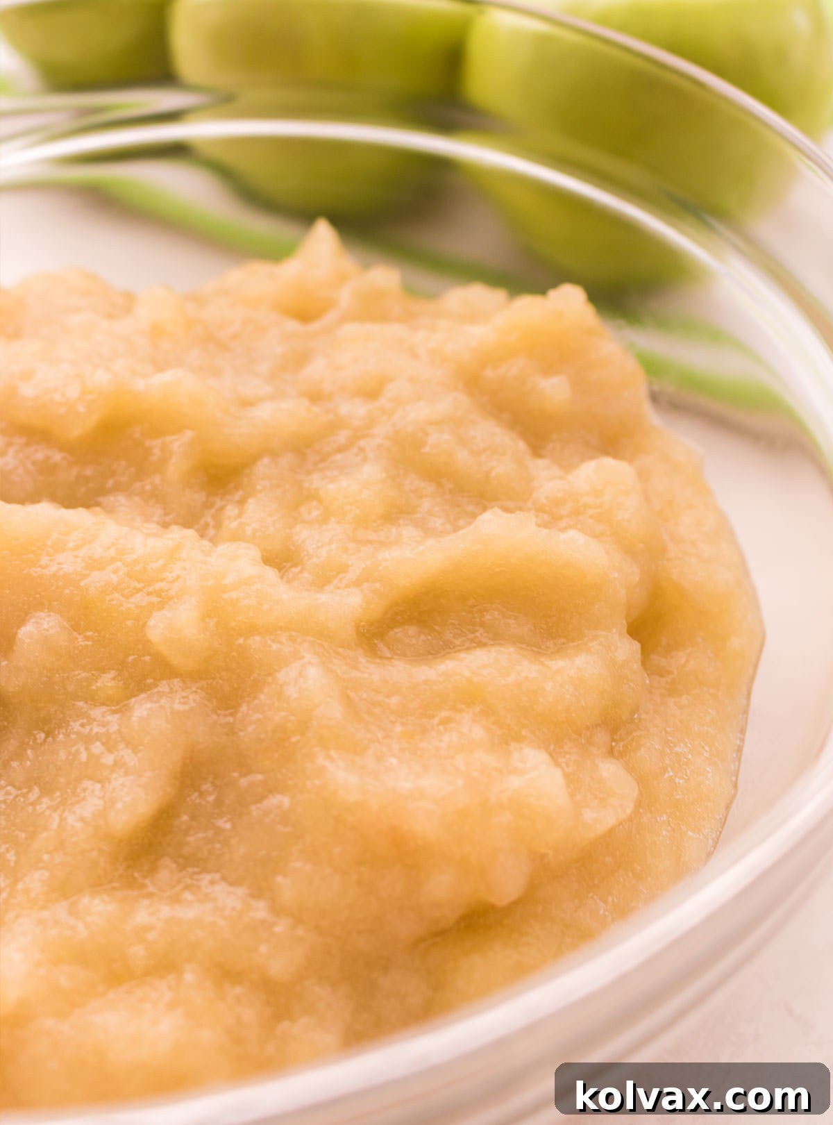 Closeup on a glass bowl filled with The Best Homemade Applesauce sitting on a white table in front of green apples, with a spoon resting in the bowl.