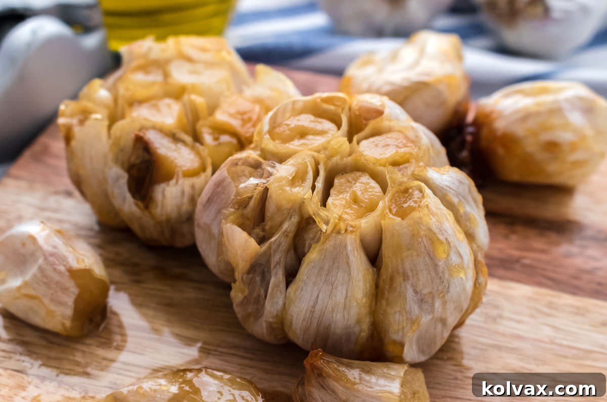 Heads of roasted garlic sitting on a cutting board in front of a blue and white towel, olive oil and a garlic press.