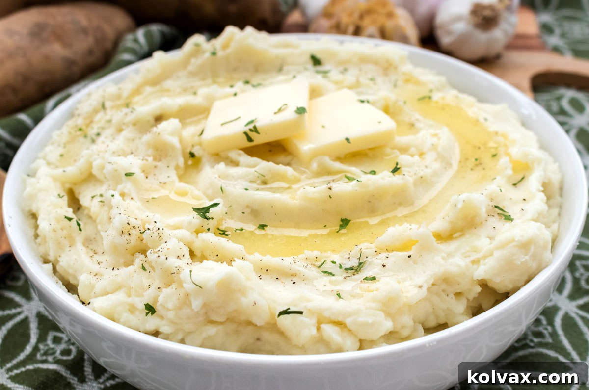 Closeup on a white serving bowl filled with Roasted Garlic Mashed Potatoes sitting on a green towel with potatoes and garlic in the background.