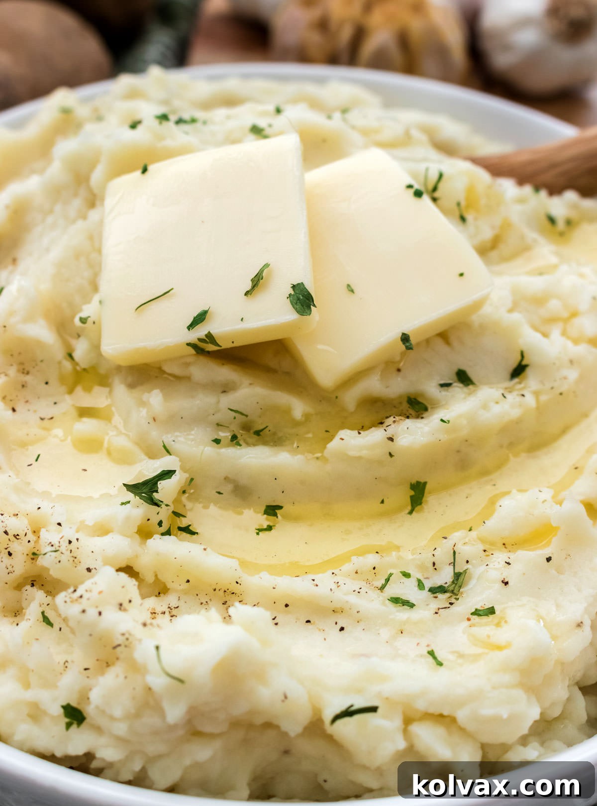 Closeup on a white serving bowl filled with Roasted Garlic Mashed Potatoes topped with two pats of butter and parsley.