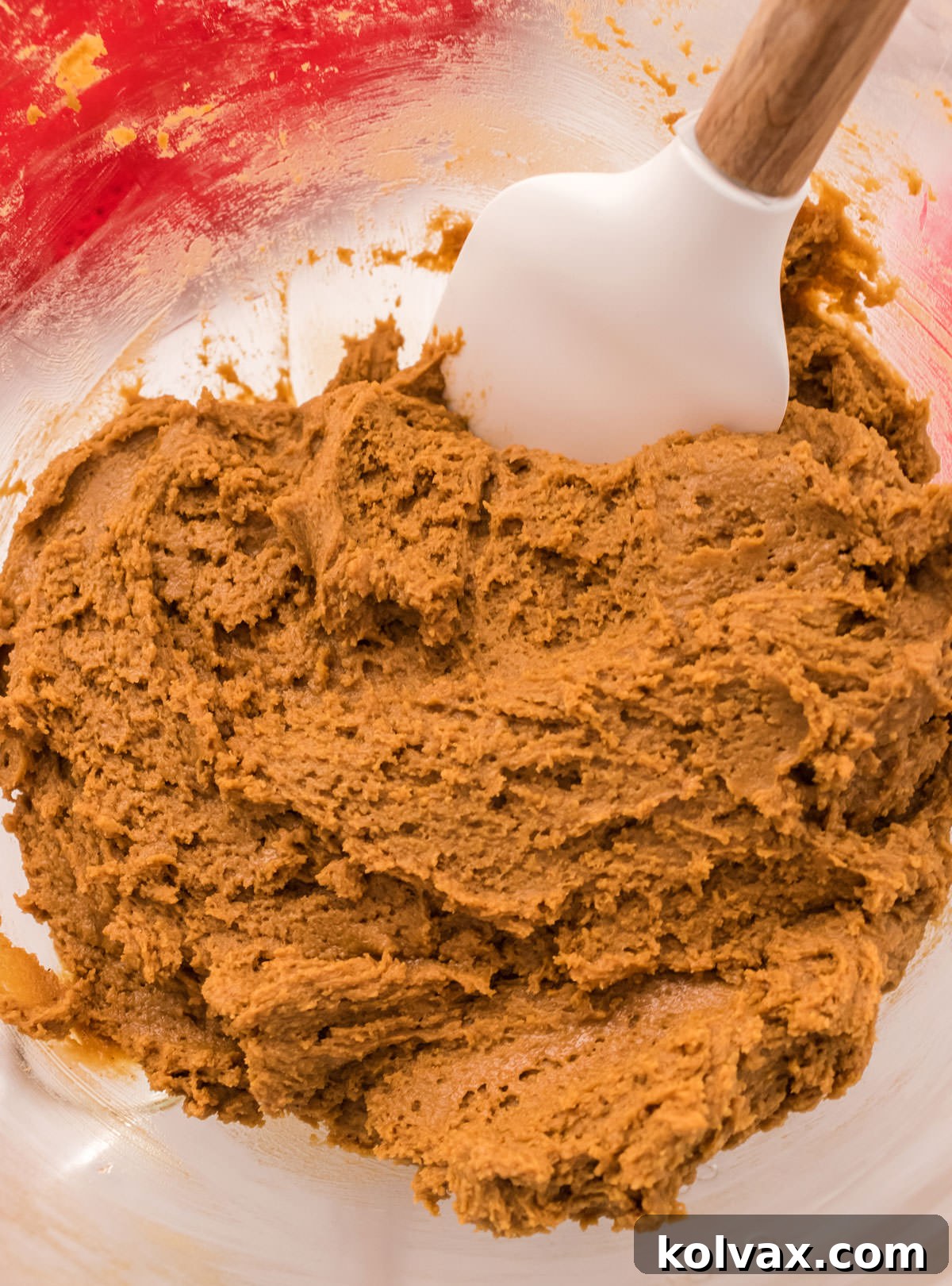 A close-up view of the rich, brown Soft Molasses Cookie dough resting in a clear glass mixing bowl, with a white spatula visible at the edge, ready for shaping.