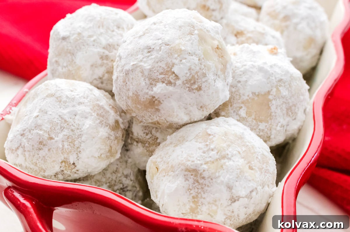 Close up of a bunch of Snowball Cookies in a container sitting on a red kitchen towel. The cookies are round, covered in white powdered sugar, and some visible pecan pieces. They are arranged appealingly in a festive red container.