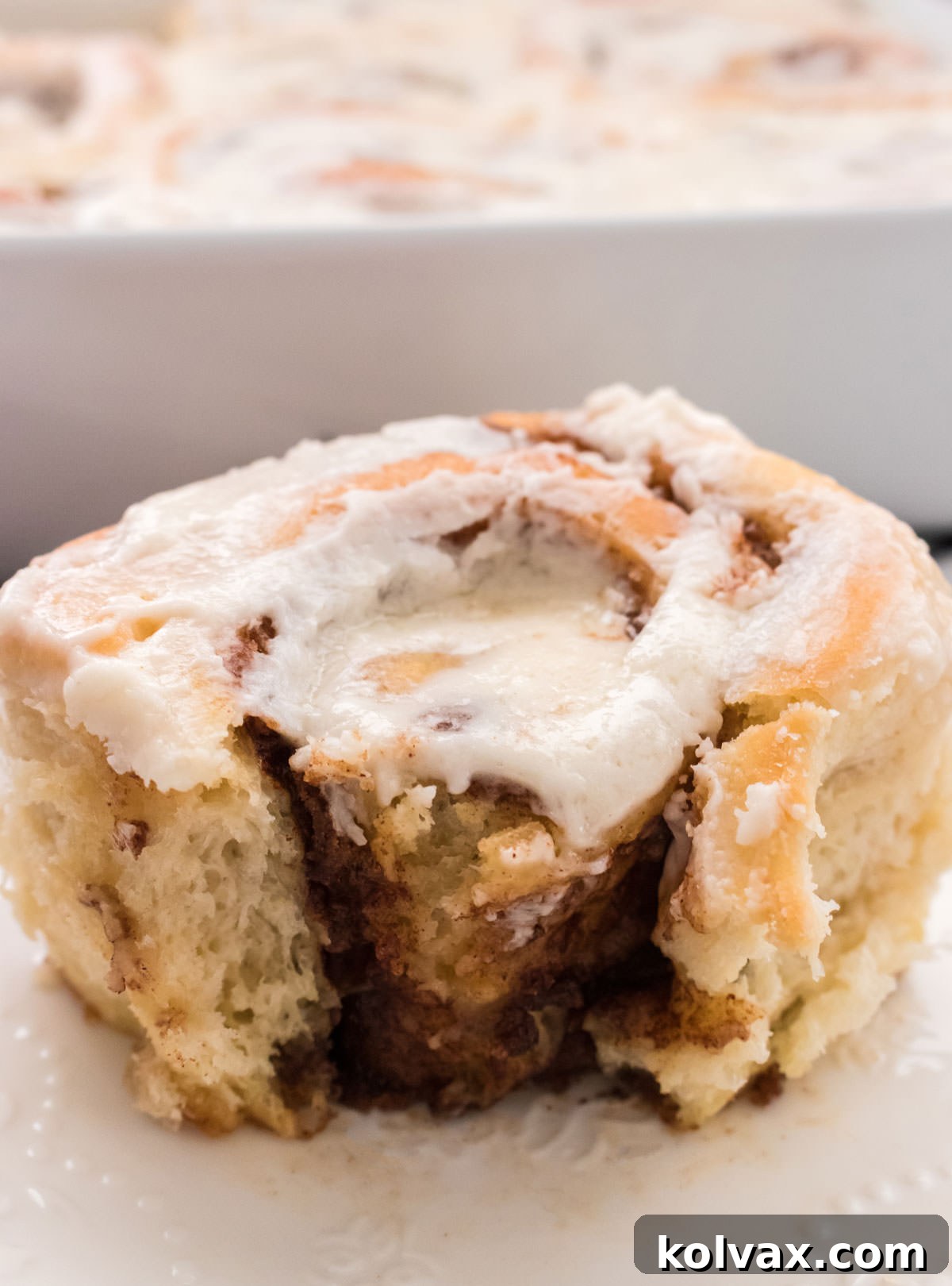 A homemade cinnamon roll with a bite taken out, sitting on a white plate in front of a baking pan full of other freshly baked cinnamon rolls, all frosted and ready to eat.
