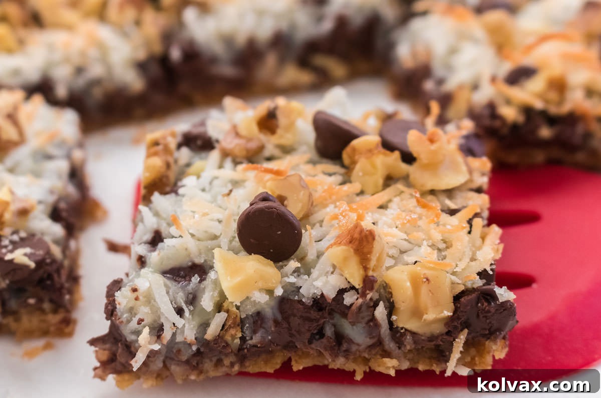 Close up of a single Magic Bar with golden brown top and visible layers, sitting in front of a pan of freshly baked Magic Bars.