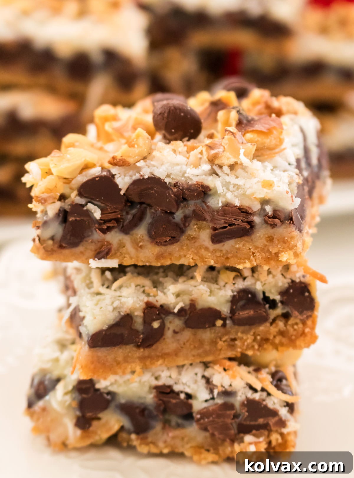 Close up on a neat stack of three Magic Bars, showing their distinct layers and gooey texture, positioned in front of a blurred background of more bars.